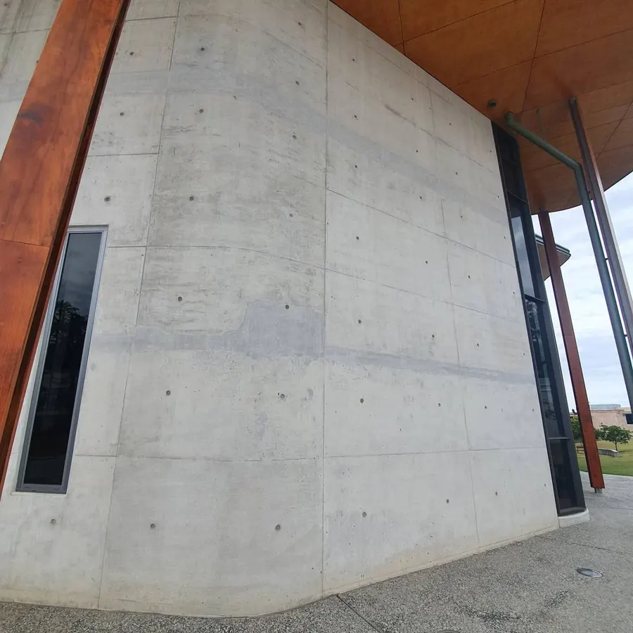 A curved off-form concrete wall with tie holes and panel lines under a timber soffit