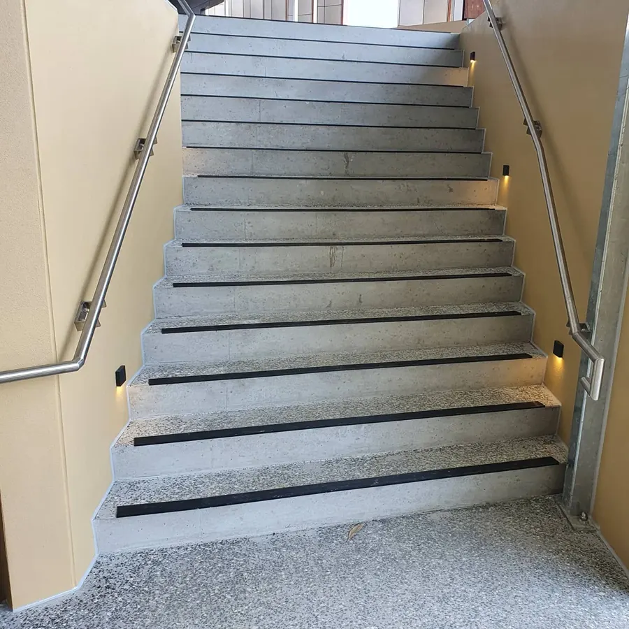 A concrete stair with black nosing strips between rendered walls and steel handrails